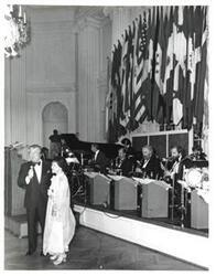 ["Black and white photograph of Mary Albert apparently being introduced at the American Revolution Bicentennial Ball in Oklahoma. She is standing beside a man with a microphone. A display of flags and an orchestra is shown behind them. October 02, 1976."]