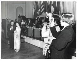 ["Black and white photograph of Mary Albert apparently being introduced at the American Revolution Bicentennial Ball in Oklahoma. She is standing beside a man with a microphone. A display of flags and an orchestra is shown behind them. October 02, 1976."]