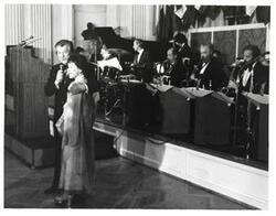 ["Black and white photograph of Mary Albert apparently being introduced at the American Revolution Bicentennial Ball in Oklahoma. She is standing beside a man with a microphone. A display of flags and an orchestra is shown behind them. October 02, 1976."]
