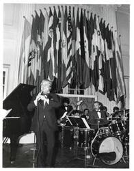 ["Black and white photograph of Unidentified man holding a microphone and standing in front of the band at the American Revolution Bicentennial Ball in Oklahoma. October 2, 1976"]