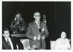 ["Black and white photograph print of Robert Michel speaking into a microphone."]