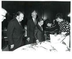 ["Carl Albert (center), Alan Cranston (behind Albert), and Roz Wyman looking at a scrapbook/photo album at a luncheon."]
