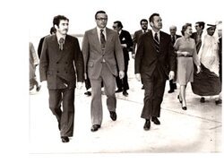 ["The image depicts Dewey Bartlett walking with Fawaz Kudsi, Charge Phillip Griffin, Robert Smith, Nathaniel Howell, Jr., Mrs. Hansen, John Limbert and Rahma Masood across an airstrip"]
