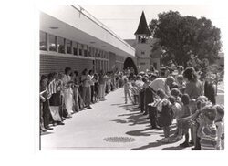 ["The image depicts Dewey Bartlett shaking hands with many school children at the Medford Public School"]